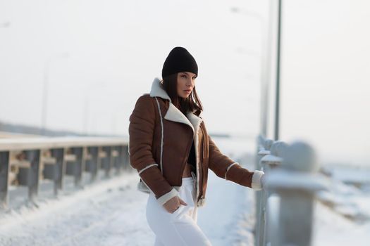 Alluring woman wearing brown sheepskin jacket with hat and looking at camera standing on snowy city bridge