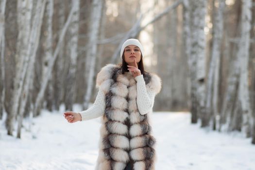 Gorgeous young brunette in fur vest and white knitwear looking confidently away against trees in winter woods