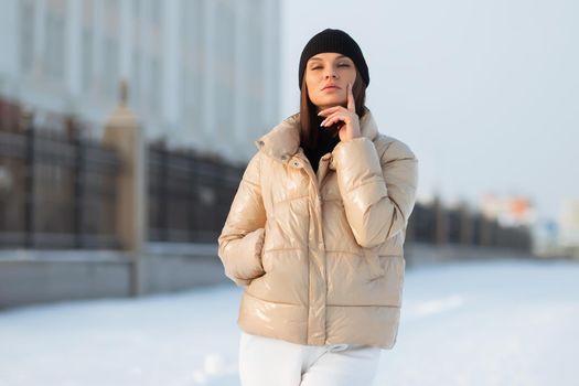 Alluring woman wearing brown sheepskin jacket with hat and looking at camera standing on snowy city bridge