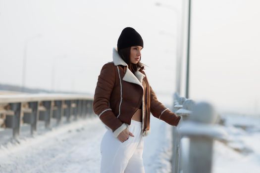 Alluring woman wearing brown sheepskin jacket with hat and looking at camera standing on snowy city bridge