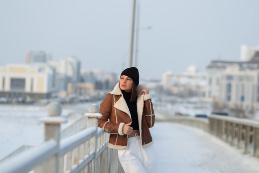 Alluring woman wearing brown sheepskin jacket with hat and looking at camera standing on snowy city bridge