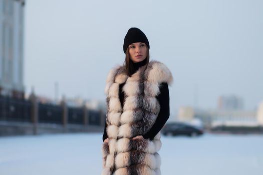 Alluring woman wearing brown sheepskin jacket with hat and looking at camera standing on snowy city bridge