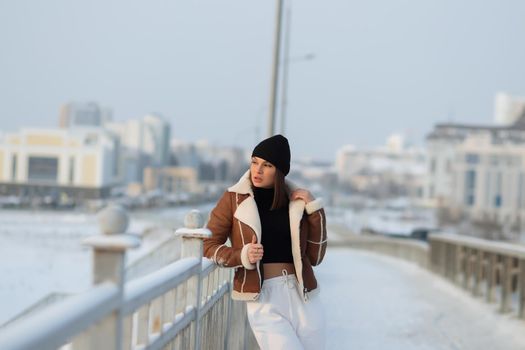 Alluring woman wearing brown sheepskin jacket with hat and looking at camera standing on snowy city bridge