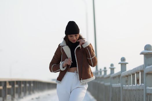 Alluring woman wearing brown sheepskin jacket with hat and looking at camera standing on snowy city bridge