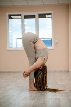 Young woman doing yoga in the gym. A girl with a long braid and in a beige tracksuit stands in a stork pose on a pink carpet. A woman performs padahstasana