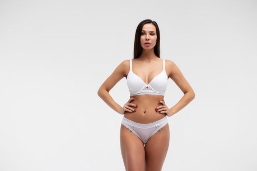 Confident female wearing white underwear standing with hands on waist on white background and looking at camera