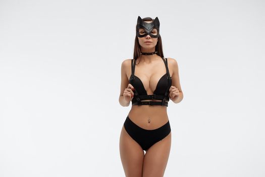 Slim seductive female wearing black mask and sexy underwear standing with hands on waist on white background and looking at camera