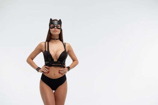 Slim seductive female wearing black mask and sexy underwear standing with hands on waist on white background and looking at camera