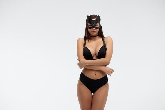 Slim seductive female wearing black mask and sexy underwear standing with hands on waist on white background and looking at camera