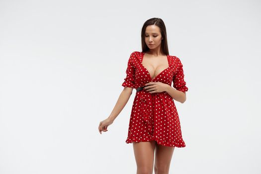 Sexy female wearing red short polka dot dress standing on white background in studio and touching hair while looking at camera