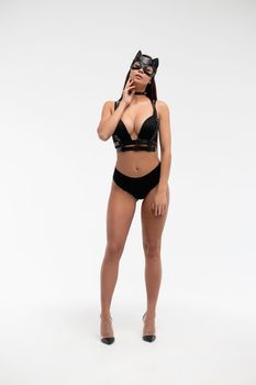 Slim seductive female wearing black mask and sexy underwear standing with hands on waist on white background and looking at camera