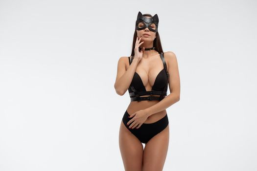 Slim seductive female wearing black mask and sexy underwear standing with hands on waist on white background and looking at camera