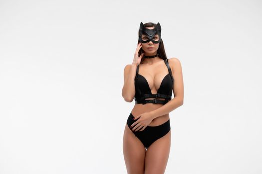Slim seductive female wearing black mask and sexy underwear standing with hands on waist on white background and looking at camera