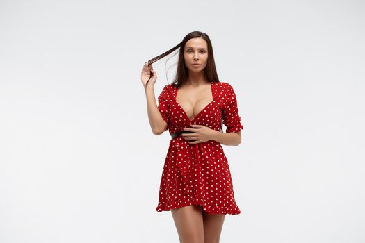 Sexy female wearing red short polka dot dress standing on white background in studio and touching hair while looking at camera