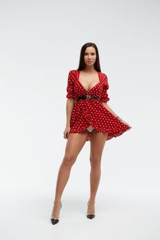 Sexy female wearing red short polka dot dress standing on white background in studio and touching hair while looking at camera