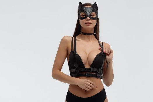 Slim seductive female wearing black mask and sexy underwear standing with hands on waist on white background and looking at camera