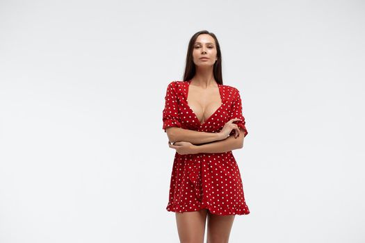 Sexy female wearing red short polka dot dress standing on white background in studio and touching hair while looking at camera