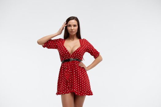 Sexy female wearing red short polka dot dress standing on white background in studio and touching hair while looking at camera