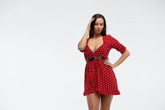 Sexy female wearing red short polka dot dress standing on white background in studio and touching hair while looking at camera