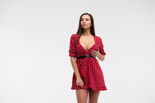 Sexy female wearing red short polka dot dress standing on white background in studio and touching hair while looking at camera