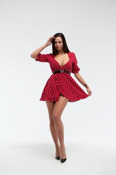 Sexy female wearing red short polka dot dress standing on white background in studio and touching hair while looking at camera