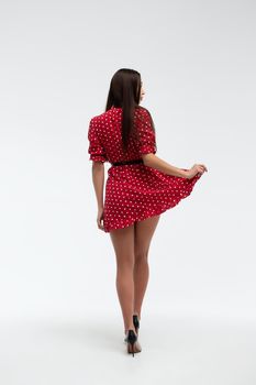 Sexy female wearing red short polka dot dress standing on white background in studio and touching hair while looking at camera