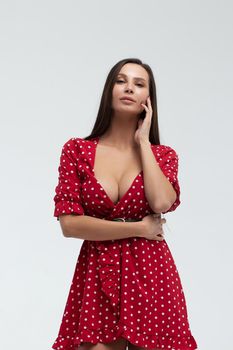 Sexy female wearing red short polka dot dress standing on white background in studio and touching hair while looking at camera