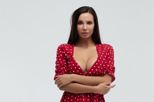 Sexy female wearing red short polka dot dress standing on white background in studio and touching hair while looking at camera