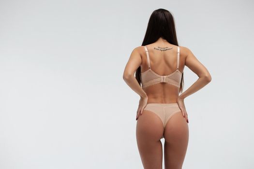 Back view of unrecognizable slim female wearing beige lingerie standing on white background in studio