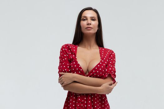 Sexy female wearing red short polka dot dress standing on white background in studio and touching hair while looking at camera