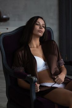 Sensual young female with long dark hair in stylish dress with decollete relaxing in comfortable armchair and looking away