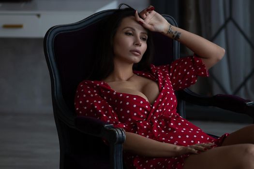 Sensual young female with long dark hair in stylish dress with decollete relaxing in comfortable armchair and looking away