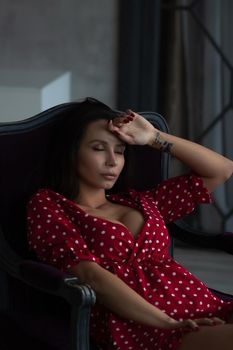 Sensual young female with long dark hair in stylish dress with decollete relaxing in comfortable armchair and looking away