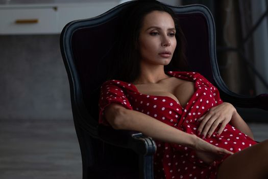 Sensual young female with long dark hair in stylish dress with decollete relaxing in comfortable armchair and looking away