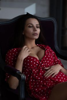 Sensual young female with long dark hair in stylish dress with decollete relaxing in comfortable armchair and looking away