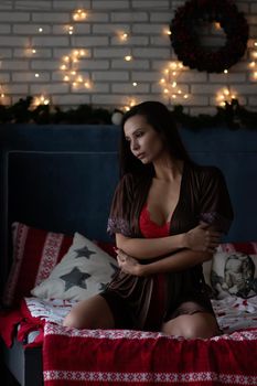 Self assured young lady in brown silk robe and white bra sitting on comfortable bed in cozy room decorated with glowing garlands and wreath during Christmas holidays