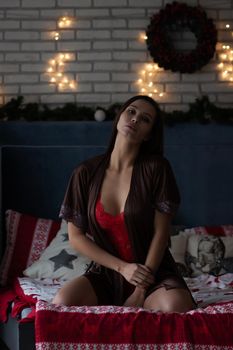 Self assured young lady in brown silk robe and red bodysuit sitting on comfortable bed in cozy room decorated with glowing garlands and wreath during Christmas holidays