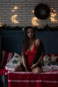 Self assured young lady in brown silk robe and red bodysuit sitting on comfortable bed in cozy room decorated with glowing garlands and wreath during Christmas holidays