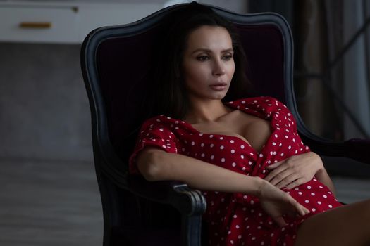 Sensual young female with long dark hair in stylish dress with decollete relaxing in comfortable armchair and looking away