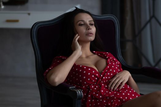 Sensual young female with long dark hair in stylish dress with decollete relaxing in comfortable armchair and looking away