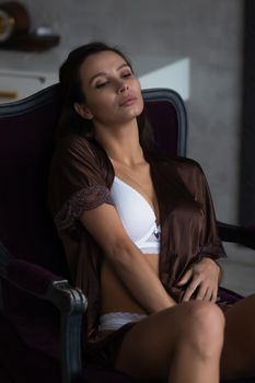 Sensual young female with long dark hair in stylish dress with decollete relaxing in comfortable armchair and looking away