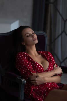 Sensual young female with long dark hair in stylish dress with decollete relaxing in comfortable armchair and looking away