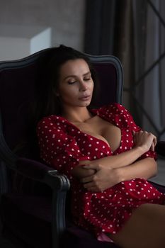 Sensual young female with long dark hair in stylish dress with decollete relaxing in comfortable armchair and looking away