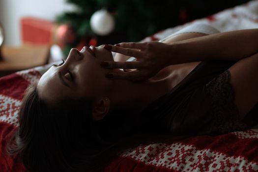 Side view of sexual young female with long dark hair in elegant red bra lying on comfortable bed with closed eyes near decorated Christmas tree