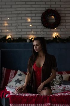 Self assured young lady in brown silk robe and red bodysuit sitting on comfortable bed in cozy room decorated with glowing garlands and wreath during Christmas holidays