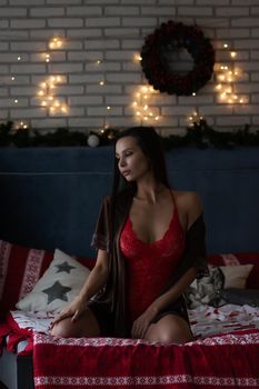 Self assured young lady in brown silk robe and red bodysuit sitting on comfortable bed in cozy room decorated with glowing garlands and wreath during Christmas holidays