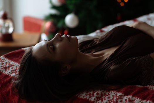 Side view of sexual young female with long dark hair in elegant red bra lying on comfortable bed with closed eyes near decorated Christmas tree