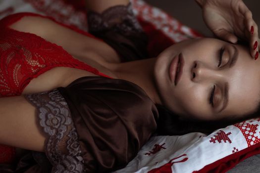 Side view of sexual young female with long dark hair in elegant red bra lying on comfortable bed with closed eyes near decorated Christmas tree