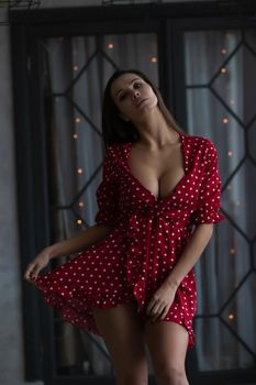 Self esteem young female with long hair in stylish mini dress with decollete standing in dark bedroom with hand on waist and looking at camera