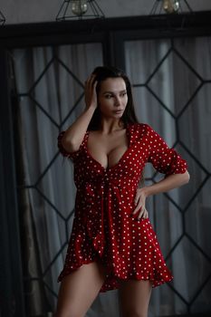Self esteem young female with long hair in stylish mini dress with decollete standing in dark bedroom with hand on waist and looking at camera